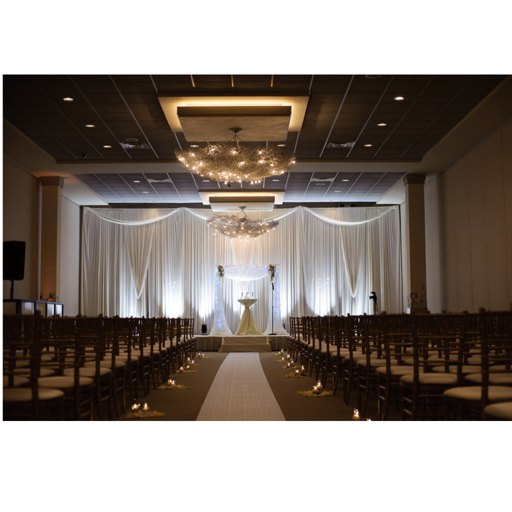 Elegant indoor wedding ceremony setup with rows of chairs facing a white aisle, flanked by tall floral arrangements on white columns, leading to a decorated altar with white drapery and modern chandeliers overhead.