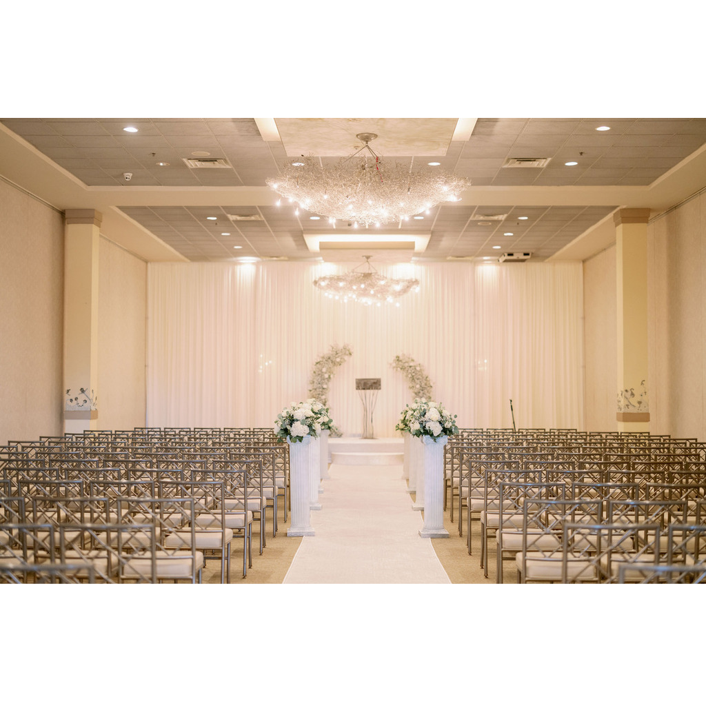 Elegant indoor wedding ceremony setup with rows of gold chairs facing a white aisle, flanked by tall floral arrangements on white columns, leading to a decorated altar with white drapery, floral arches, and modern chandeliers overhead.